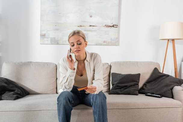 blonde woman holding credit card and talking on smartphone in living room  - Fotó, kép