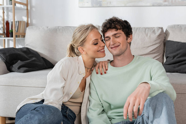 happy blonde woman with closed eyes smiling near boyfriend in living room  - Foto, afbeelding