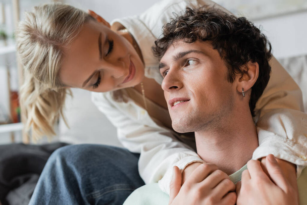smiling and blonde woman embracing curly man in living room  - Φωτογραφία, εικόνα