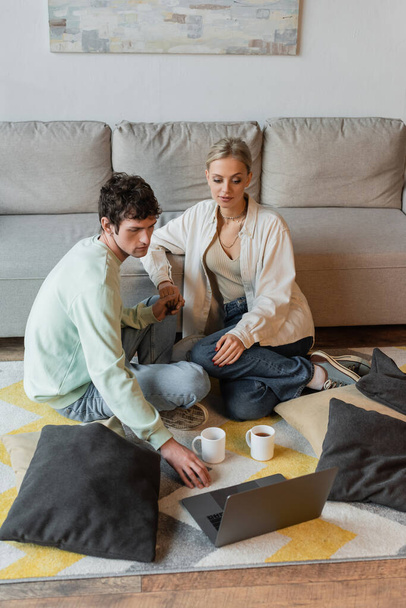 curly man holding hand of blonde woman while looking at laptop near cups on carpet  - Foto, Bild