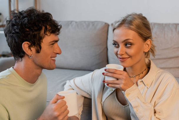 happy woman with blonde hair and curly man looking at each other while holding cups with coffee  - Zdjęcie, obraz