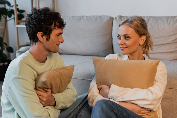 happy woman with blonde hair and curly man looking at each other while holding pillows  - Fotoğraf, Görsel
