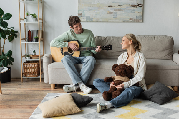 curly man sitting on couch and playing acoustic guitar near happy woman with teddy bear  - Foto, immagini