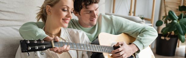 curly man playing acoustic guitar near cheerful blonde woman while sitting near couch, banner  - Foto, immagini