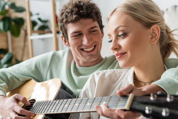 happy man playing acoustic guitar near blonde woman on blurred foreground  - Photo, Image
