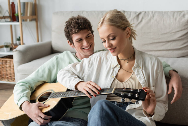 curly man playing acoustic guitar near pleased blonde woman while sitting in living room  - Foto, imagen
