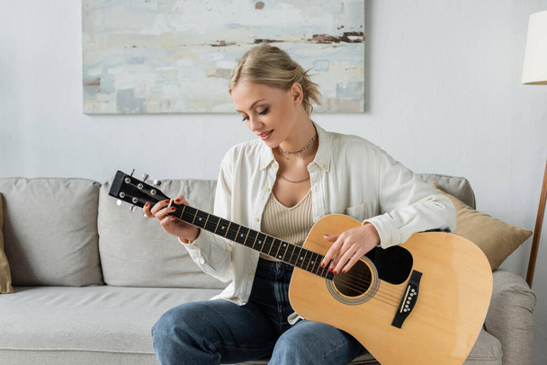 cheerful blonde woman playing acoustic guitar while sitting on couch  - Fotó, kép