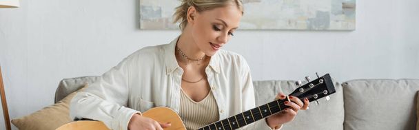 cheerful blonde woman playing acoustic guitar in living room, banner  - 写真・画像