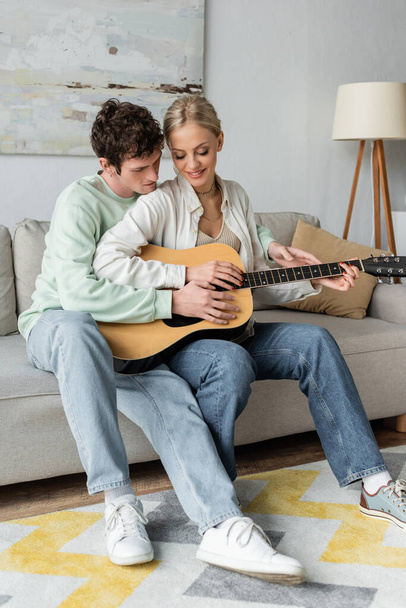 happy blonde woman playing acoustic guitar with curly boyfriend while sitting on couch  - 写真・画像