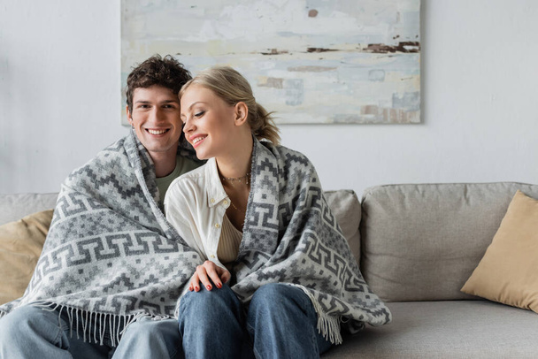 happy young man and woman covered in blanket sitting on couch  - Fotoğraf, Görsel