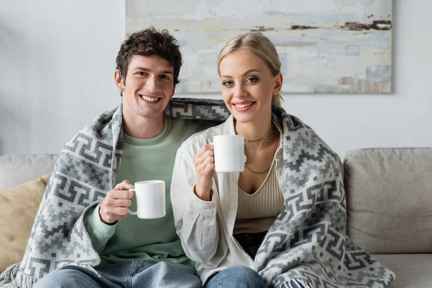 happy young man and smiling blonde woman covered in blanket holding cups of tea while sitting on couch  - Fotó, kép
