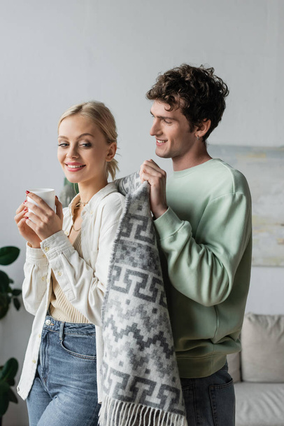 cheerful and caring man covering smiling blonde woman with blanket  - Foto, Imagen