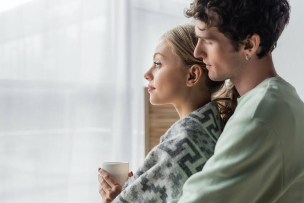 side view of caring man hugging blonde girlfriend covered in blanket and holding cup of tea  - Foto, Bild