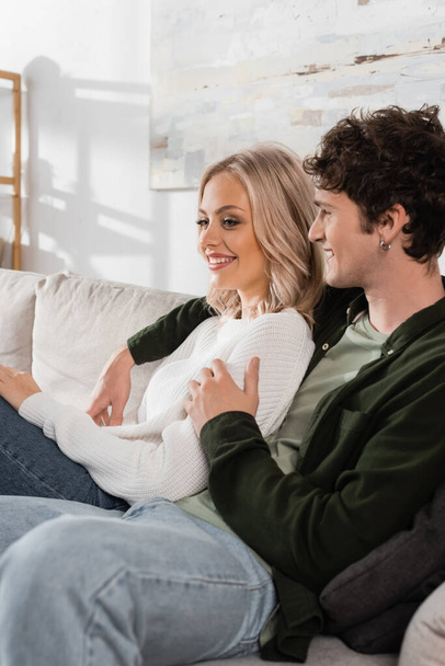 smiling and curly man in shirt hugging pretty blonde girlfriend in white sweater  - Fotó, kép