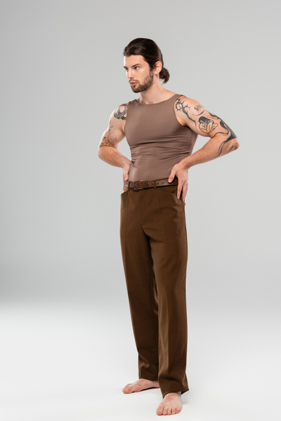 Barefoot and tattooed man in pants and tank top posing on grey background - Foto, imagen