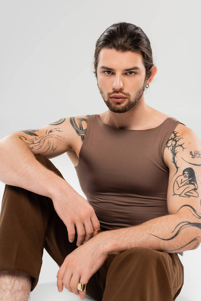 Stylish tattooed man in sleeveless short sitting on grey background - Photo, Image