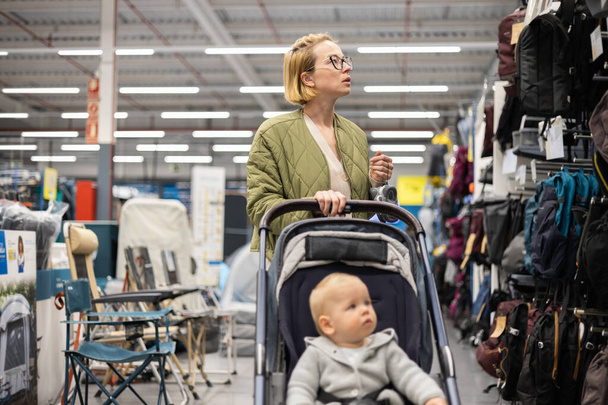 Casualy dressed mother choosing sporty shoes and clothes products in sports department of supermarket store with her infant baby boy child in stroller - Foto, Imagen