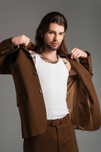 Trendy long haired man with tattoo wearing jacket isolated on grey  - Foto, imagen