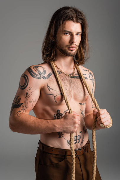 Sexy tattooed model posing with rope and looking at camera isolated on grey  - Valokuva, kuva