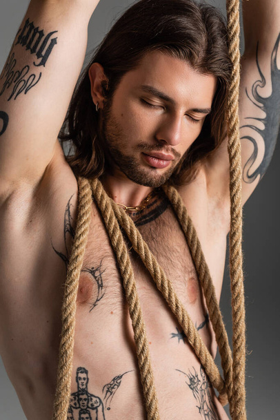 Long haired and tattooed model posing with rope isolated on grey  - Фото, изображение