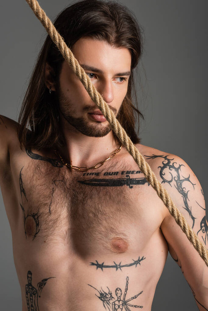 Shirtless tattooed man looking away near rope isolated on grey  - 写真・画像