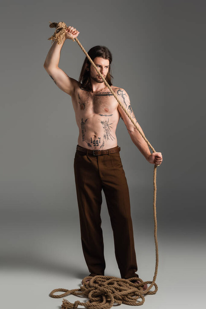 Full length of tattooed model in pants holding rope on grey background  - 写真・画像