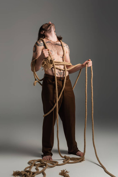 Log haired and tattooed model in pants holding rope on grey background  - Foto, immagini