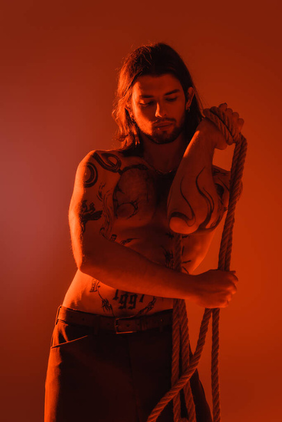 Muscular tattooed man holding rope isolated on red with light  - Φωτογραφία, εικόνα
