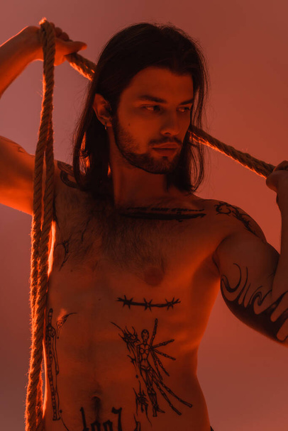 Young tattooed and shirtless model posing with rope isolated on red  - 写真・画像