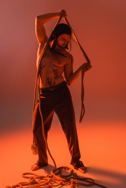 Повна довжина сексуальної татуйованої моделі позує з мотузкою на червоному тлі
  - Фото, зображення