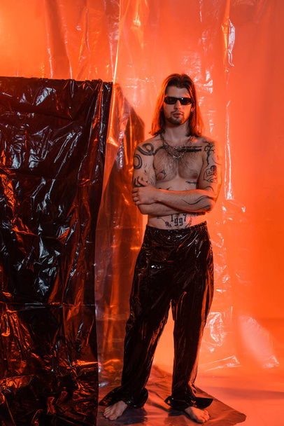 Tattooed man in latex pants and sunglasses standing near cellophane  - Photo, image