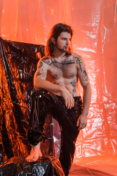 Sexy tattooed man in latex pants standing near cellophane  - Foto, imagen