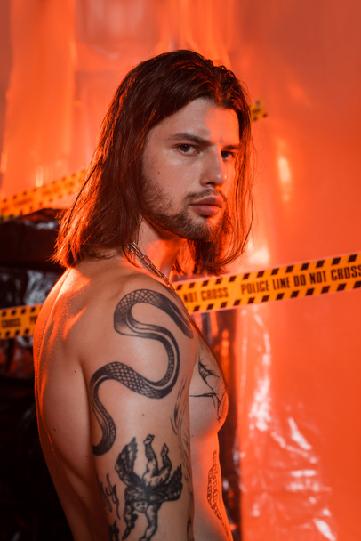 Portrait of shirtless tattooed man looking at camera near blurred police line and polyethylene - Φωτογραφία, εικόνα