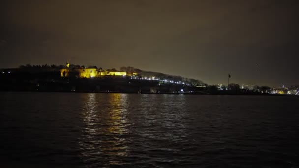The sea where the yellow lights of the city are reflected.A view of Istanbul in the summer night. Topkapi Palace view from the sea.Night sea sparkles bright light water moonlight. - Felvétel, videó