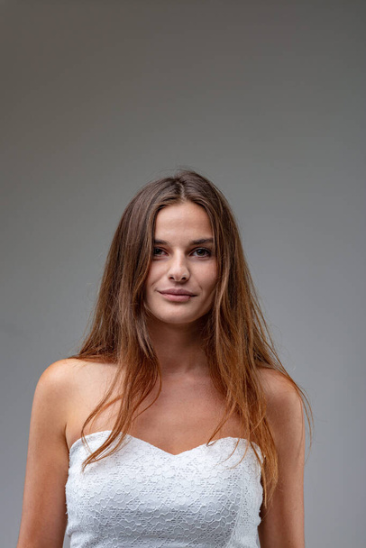 frontal half-length portrait of charming and attractive young woman with long brown hair, she wears a white sheath dress and her shoulders leave her skin bare, she smiles quietly and confidently - 写真・画像