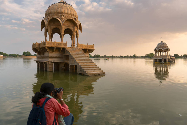 Indian female traveller, woman photographer taking picture of Chhatris and shrines with reflection of them on the water of Gadisar lake. Sun set and colorful clouds on sky. Jaisalmer, Rajasthan,India. - Фото, изображение
