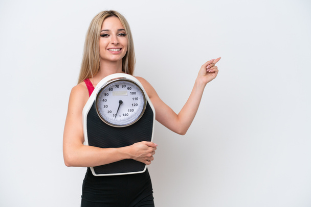 Pretty blonde woman isolated on white background with weighing machine and pointing side - Valokuva, kuva