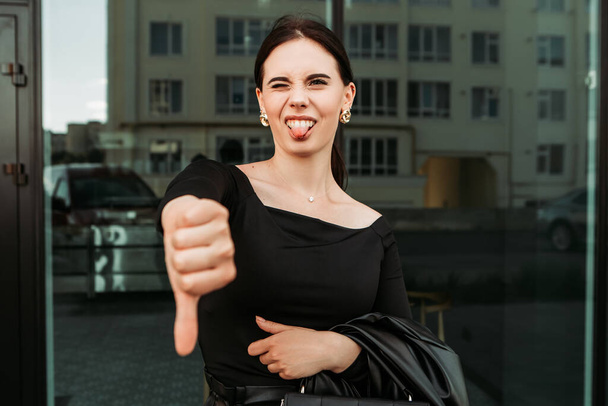 Pretty brunette caucasian woman standing outside street with white teeth beaming healthy lady. Happy millennial girl showing thumb down dislike disapprove sign gesturing making faces show tongue - Photo, image