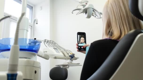 歯の椅子の若いブロンドは、スマートフォンからインターネットを介して通信します。入院中の歯科治療前の女性患者. - 映像、動画