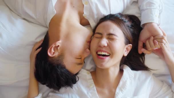 Top view of true love young asia couple lying down kiss wife face cheek on cozy bed. Relax awake morning fall in love of just married sweet lover asian people joy laugh toothy smile at home bedroom. - Séquence, vidéo