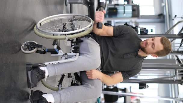 Vertical video of young attractive smiling man in wheelchair lifting dumbbells in gym. Rehabilitation of people with disabilities - Filmati, video