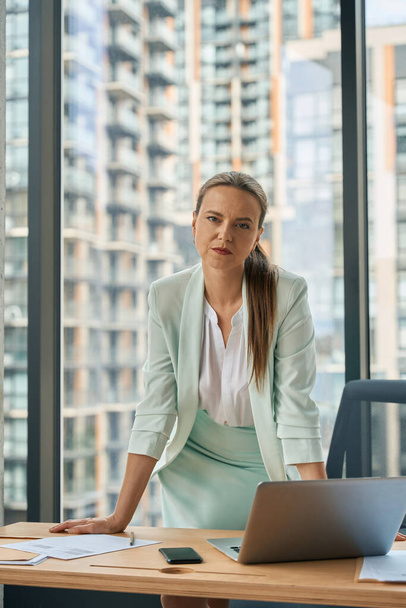 Tired young woman in business-like outfit leaning on her office desk and strictly looking at the camera - Foto, Bild