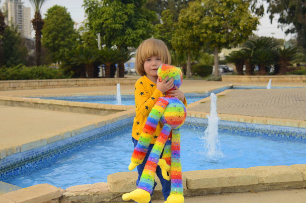 Ένα θετικό μικρό αγόρι, 4 ετών, περπατάει στο πάρκο με ένα φωτεινό χρωματιστό παιχνίδι. Concept: νηπιαγωγείο, φανταστικό φίλο, αγαπημένο παιχνίδι. - Φωτογραφία, εικόνα