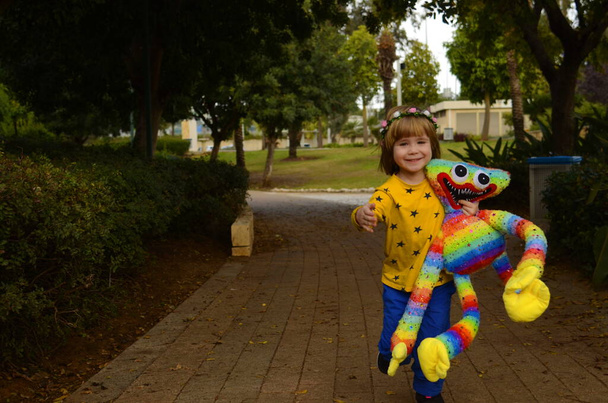 A cute boy in a yellow t-shirt runs along the path in the park with a large multi-colored soft toy. The child received a gift, the kid rejoices at a new friend. Concept: children and monsters, scary toys, imaginary friends. - Foto, Imagem