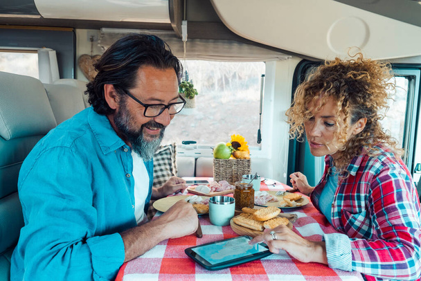 Couple man and woman inside a camper van planning the next travel destination together using GPS modern device on the table. Lunchtime on vehicle motor home alternative adventure vacation. Lifestyle - Foto, imagen