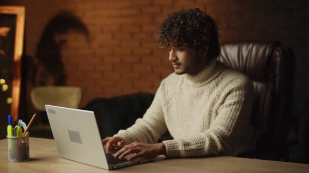 Curly young man sending message in a work chat and smiling. Freelancer in sweater typing on laptop keyword in his home office. Work from home concept. High quality 4k footage - Materiał filmowy, wideo