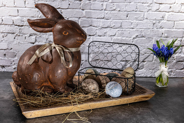 brown clay easter bunny in wooden bowl with easter eggs and hay in front of white brick wall - Foto, Imagen