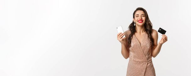 Online shopping and holidays concept. Satisfied and happy woman in elegant dress smiling, using credit card and mobile phone, standing over white background. - Photo, image