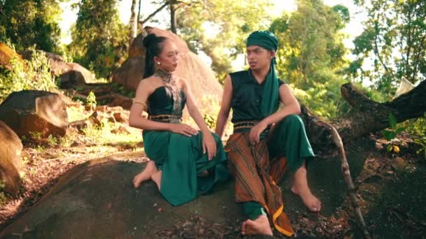 An Asian couple flirting on the top of the rock while sitting together in green clothes with the forest in the background during the daylight - Video