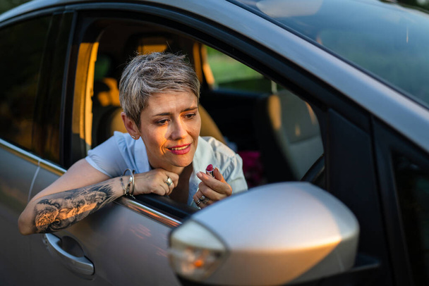 One woman mature caucasian female businesswoman sitting in car putting lipstick fixing repairing makeup on her face while waiting in summer day evening real people copy space gray hair - Φωτογραφία, εικόνα